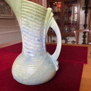 Könnte beinhalten: Ein Keramikkrug mit bauchiger Basis, ausgestelltem Ausguss und einem Henkel. Die Oberfläche zeigt eine gesprenkelte Glasur in blassen Blau-, Creme- und sanften Gelbtönen mit horizontalen Rillen, die sich um den Körper und den Hals ziehen.