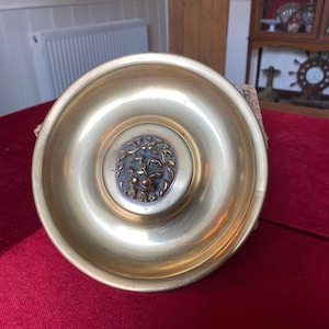 May include: A circular brass door knocker featuring a polished gold-toned finish and a central medallion with a detailed, raised relief design of a lion head. The metal surface is smooth and reflective, showcasing a classic, rounded shape.
