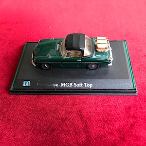 Może przedstawiać: Ciemnozielony model w skali 1:43 samochodu MGB Soft Top z otwieranym dachem. Model ma czarny dach, srebrne akcenty i stoi na czarnej podstawie ekspozycyjnej z zieloną obwódką. Na podstawie wydrukowano napis "1/43 MGB Soft Top".