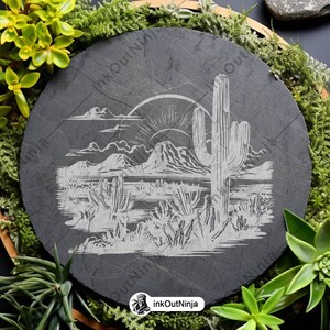 May include: A black slate coaster with a white etched design of a desert landscape with a large cactus and a rising sun. The coaster is surrounded by green plants.