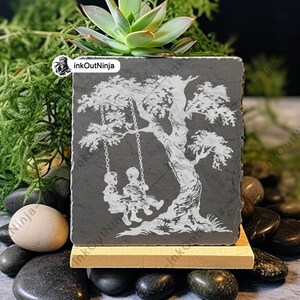 May include: A gray slate coaster with a white silhouette of two children on a swing set under a tree. The coaster is on a wooden stand and surrounded by small gray and brown stones.