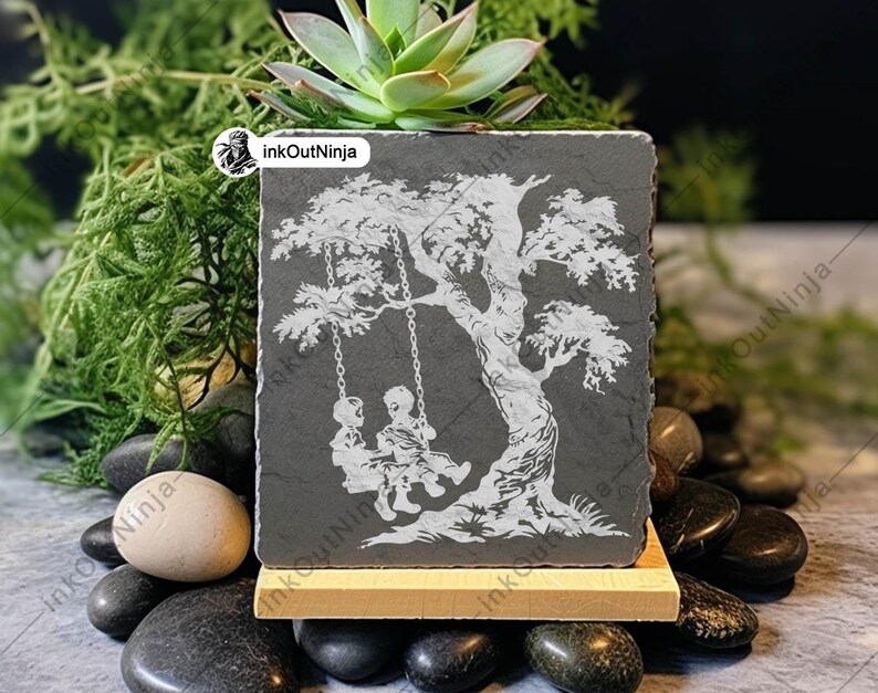 May include: A gray slate coaster with a white silhouette of two children on a swing set under a tree. The coaster is on a wooden stand and surrounded by small gray and brown stones.
