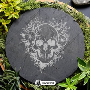 May include: A black slate coaster with a white engraved design of a skull with flowers. The coaster is round and has a rustic look.
