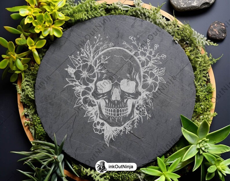 May include: A black slate coaster with a white engraved design of a skull with flowers. The coaster is round and has a rustic look.