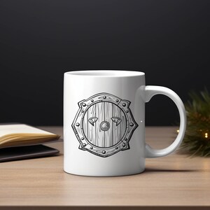 May include: White ceramic mug with a black and white illustration of a shield with a wooden texture. The shield has a circular center with a small circle in the middle and two small triangles on either side.