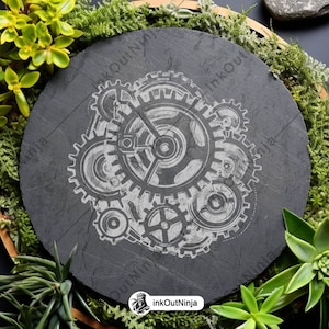 May include: A black slate coaster with a white design of interlocking gears. The coaster is surrounded by green moss and succulents.
