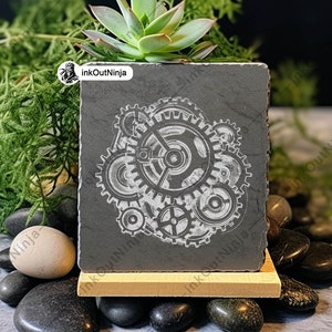 May include: A black slate square planter with a white design of interlocking gears. The planter is sitting on a wooden stand and surrounded by small black and gray stones.
