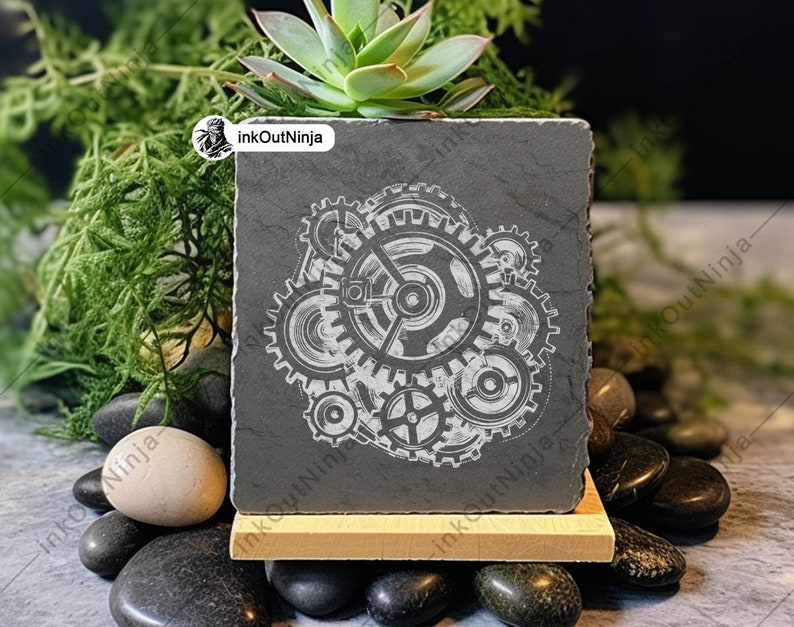 May include: A black slate square planter with a white design of interlocking gears. The planter is sitting on a wooden stand and surrounded by small black and gray stones.