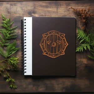 May include: A brown spiral bound notebook with a black cover. The cover has a brown outline of a shield with a wooden texture. The shield has two eyes drawn on it.