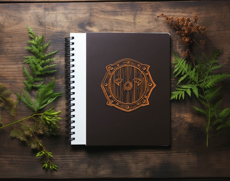 May include: A brown spiral bound notebook with a black cover. The cover has a brown outline of a shield with a wooden texture. The shield has two eyes drawn on it.