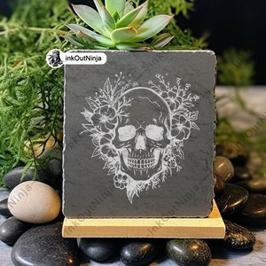 May include: A black slate square planter with a white engraved skull design surrounded by flowers. The planter is sitting on a wooden stand and surrounded by black stones.