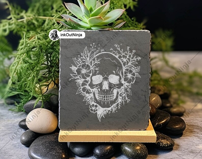 May include: A black slate square planter with a white engraved skull design surrounded by flowers. The planter is sitting on a wooden stand and surrounded by black stones.
