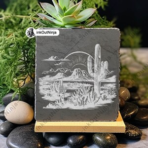 May include: A black slate coaster with a white etched design of a desert landscape with a large cactus and a mountain range in the background. The sun is setting behind the mountains. The coaster is on a wooden stand and surrounded by small black stones.