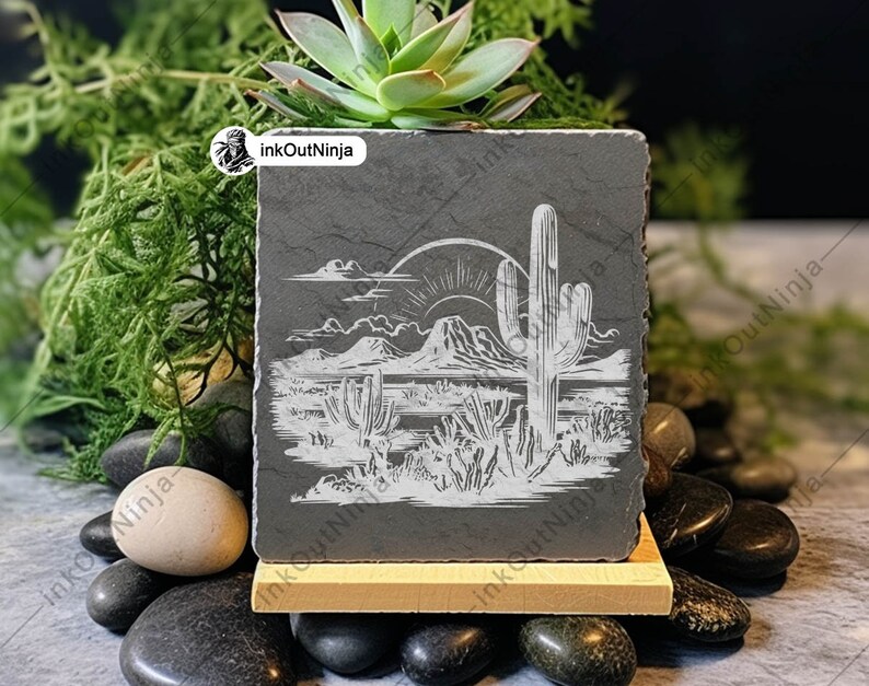 May include: A black slate coaster with a white etched design of a desert landscape with a large cactus and a mountain range in the background. The sun is setting behind the mountains. The coaster is on a wooden stand and surrounded by small black stones.