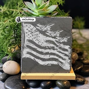 May include: A gray slate coaster with a white design of an American flag and fighter jets flying over it. The coaster is on a wooden stand and surrounded by small black and white stones.