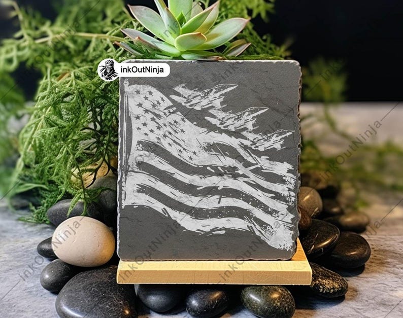 May include: A gray slate coaster with a white design of an American flag and fighter jets flying over it. The coaster is on a wooden stand and surrounded by small black and white stones.