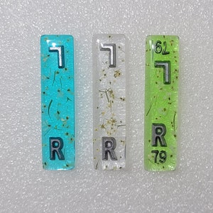 Puede incluir: Tres azulejos rectangulares con el número "7" impreso en ellos. Los azulejos están hechos de resina transparente con purpurina y pequeñas flores secas incrustadas. Los azulejos son de diferentes colores: azul, blanco y verde.