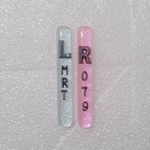 Puede incluir: Dos piezas rectangulares de resina translúcida. Una es azul claro con las letras "LMRT" en gris oscuro. La otra es rosa con los números "R079" en gris oscuro. Las piezas están sobre una superficie blanca texturizada.