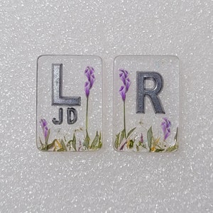 May include: Two clear resin tiles, one with a silver "L" and "JD", the other with a silver "R". Both tiles feature purple flowers and greenery. These tiles could be decorative or functional, perhaps for a craft project.