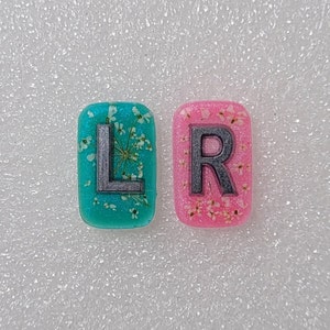 May include: Two rectangular resin charms featuring embedded dried white flowers and glitter. The left charm is teal with a dark grey letter L, and the right charm is pink with a dark grey letter R. Both pieces have rounded corners and a smooth finish.