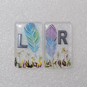 Op de afbeelding: Twee transparante harstegels met een zilveren letter, L en R, en een kleurrijk verenontwerp. De tegels zijn gevuld met gedroogde bloemen en groen.