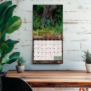May include: A wall calendar featuring a camouflage-clad figure in a forest setting. The calendar displays the month of March with a wooden border. The calendar is hanging on a white brick wall.
