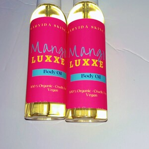 May include: Two clear glass spray bottles of body oil. The bottles have a pink label with the words "Mango Luxxe Body Oil" in yellow and blue. The label also states "100% Organic, Cruelty Free, Vegan."