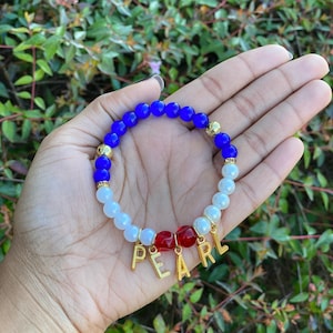 May include: A blue, white, and red beaded bracelet with gold letters spelling "PEARL".