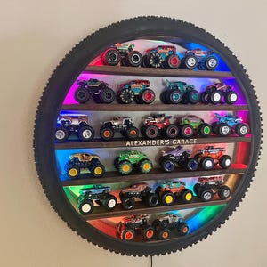 Peut inclure: Une étagère en forme de pneu avec un cadre en bois et des lumières LED arc-en-ciel présente une collection de voitures de monster truck miniatures. L'étagère est étiquetée "Alexander's Garage".
