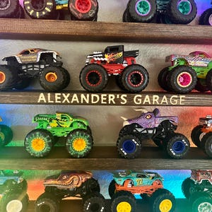 May include: A wooden shelf with a sign that reads "ALEXANDER'S GARAGE" displays a collection of monster trucks. The trucks are in various colors, including green, purple, red, and black. The trucks have large wheels and are decorated with different designs.