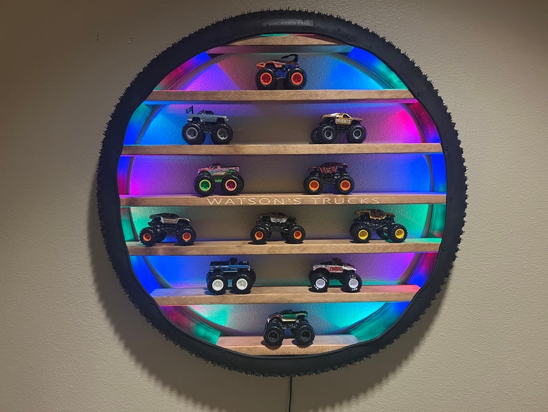 May include: A circular wooden shelf with a tire rim frame and colorful LED lights. The shelf has multiple levels and displays a collection of monster trucks. The text "WATSON'S TRUCKS" is printed on the shelf.