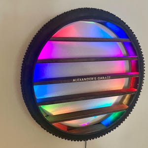 May include: A black bicycle tire rim with a rainbow LED light display and wooden shelves inside. The shelves are labeled "ALEXANDER'S GARAGE".