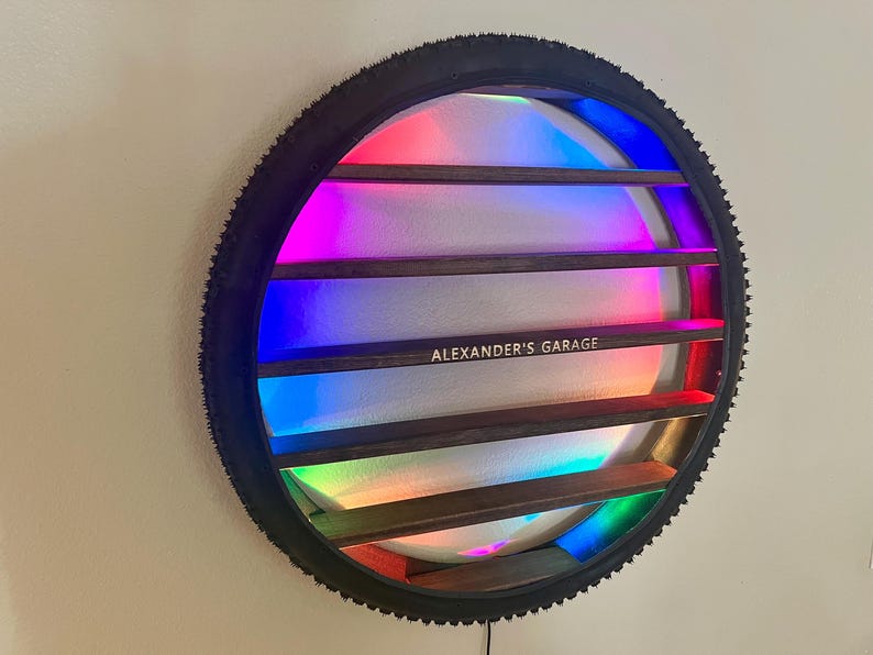 May include: A black bicycle tire rim with a rainbow LED light display and wooden shelves inside. The shelves are labeled "ALEXANDER'S GARAGE".