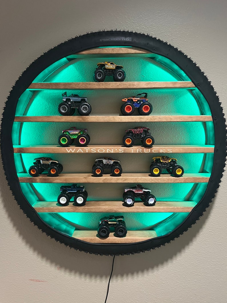 May include: A circular shelf made from a tire with wooden shelves and teal LED lights. The shelves are filled with monster trucks. The text "WATSON'S TRUCKS" is printed on the shelf.