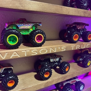 May include: A wooden shelf with a sign that reads "Watson's Trucks" displays a collection of monster trucks. The trucks are various colors, including green, red, black, and white. The trucks have different designs and logos, including "Maximum Destruction".