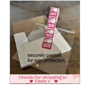 May include: A white cardboard box with a pink cardboard backing is wrapped in clear plastic. The box has a pink sticker with a cartoon elephant and the text "DO NOT CRUSH BEND OR FOLD" repeated four times. The box is labeled "securely packed for sign protection". The bottom of the image has a pink banner with the text "Thanks for shopping at Lindy's" and two pink hearts.
