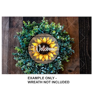 K&ouml;nnte beinhalten: Ein rundes Holzschild mit einer gemalten Sonnenblume und dem Wort "Willkommen" in wei&szlig;en Buchstaben. Das Schild ist auf einem gr&uuml;nen Kranz angebracht.