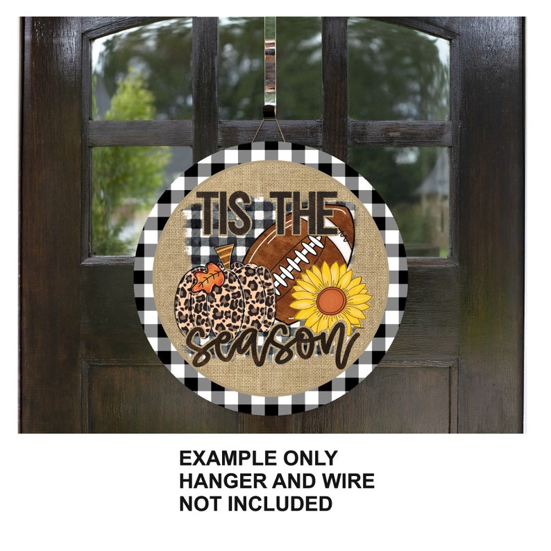 Puede incluir: Un colgador de puerta redondo con fondo a cuadros blanco y negro. El centro del colgador presenta una textura de arpillera con el texto "Tis the season" y un bal&oacute;n de f&uacute;tbol, una calabaza y un girasol.