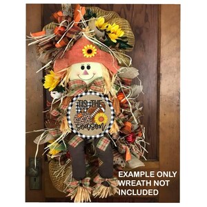 Puede incluir: Una corona de puerta de oto&ntilde;o con un espantap&aacute;jaros, girasoles y un letrero redondo que dice "Tis the season" con un bal&oacute;n de f&uacute;tbol, una calabaza y un girasol.