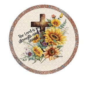 May include: A round, beige and brown glitter background with a brown wooden cross and a bouquet of sunflowers. The text "The Lord is my strength and my shield" is written in black cursive script. The text "Psalm 26:7" is written in black below the flowers.