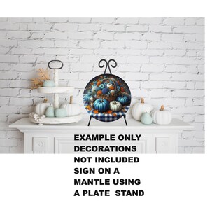 May include: A decorative plate featuring a basket of pumpkins and flowers, displayed on a black plate stand. The plate is set against a white brick wall, with additional pumpkins and a tiered tray with pumpkins. The text on the image reads: EXAMPLE ONLY, DECORATIONS NOT INCLUDED, SIGN ON A MANTLE USING A PLATE STAND.