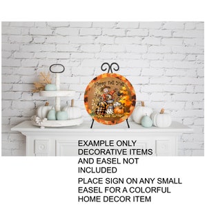 May include: A round wooden sign with a fall theme featuring a scarecrow, pumpkins, and leaves. The sign reads "Happy Fall Y'all".