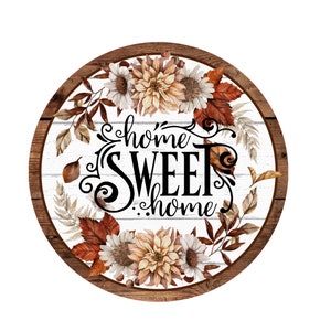 May include: Round wooden sign with a white background and a wreath of fall leaves and flowers. The sign says "home sweet home" in black lettering.