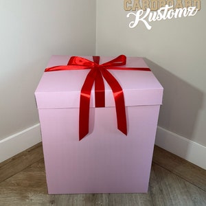 May include: A large, pink gift box with a red satin ribbon tied in a bow. The box is sitting on a wooden floor.