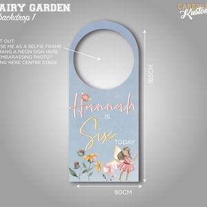 May include: A light blue door hanger with a cut-out circle in the centre. The hanger features a floral design with a fairy and the text "Hannah is Six Today". The hanger measures 18.5 cm by 8 cm.