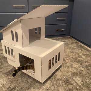 May include: A white cardboard cat house with a slanted roof and multiple openings. A cat is partially visible inside the house.