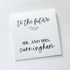 May include: A white card with the text "to the future" in a script font, with a small green leaf design. Below, it reads "MR. AND MRS. Cunningham" in a bold script font. The card is likely for a wedding or anniversary.