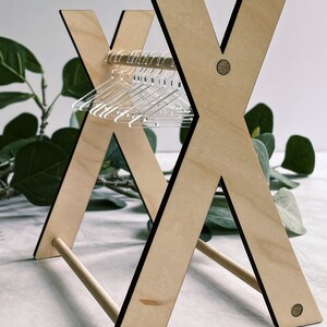 May include: A wooden clothes rack with a simple X-shaped design. The rack has a wooden dowel for hanging clothes and is made of light wood with black accents. There are clear plastic hangers on the rack.