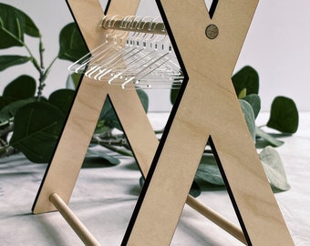 May include: A wooden clothes rack with a simple X-shaped design. The rack has a wooden dowel for hanging clothes and is made of light wood with black accents. There are clear plastic hangers on the rack.