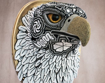 May include: A beaded eagle head sculpture with a gold beak and white and black feathers. The sculpture is mounted on a gold circular frame.